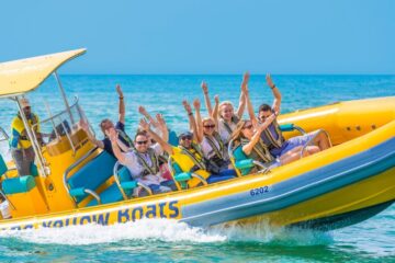 A group of cheerful passengers on a yellow speedboat raising their arms in excitement.