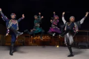 Four male dancers in vibrant, embroidered costumes leaping in unison during a traditional Middle Eastern performance.