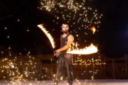 Male fire performer spinning a flaming staff amid sparkling embers.