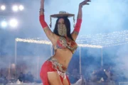 A belly dancer in a red costume with a beaded face covering, balancing a decorative prop on her head.