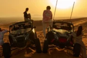 Two Can-Am buggies parked on desert dunes at sunset with four people standing nearby.