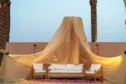 A cozy desert lounge area featuring a flowing beige canopy, white seating, and palm trees in the background.