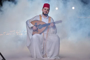 A man in traditional white attire and a red fez playing an oud amid soft desert smoke.