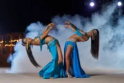 Two female dancers in matching blue costumes arching backward in a synchronized pose, enveloped by stage smoke.