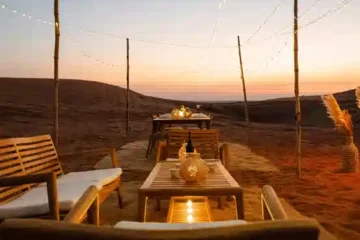 A private desert dining area at dusk, featuring simple wooden tables, soft lights, and a warm sunset backdrop.