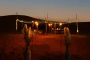 A softly lit dining setup in the desert at night, framed by small posts and hanging string lights.