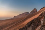 A vast sloping dune rising toward rocky hills at sunset, with tire tracks visible in the sand.