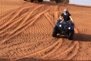 Three people riding quad bikes up a sandy dune under a bright sky.