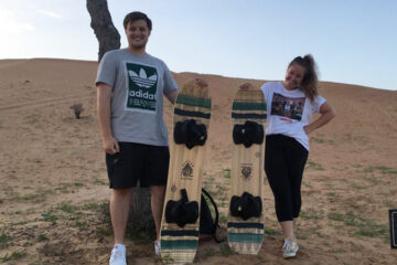 Two people holding sandboards, standing on desert ground with dunes behind them.