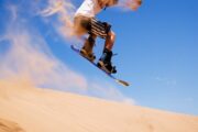 A sandboarder mid-jump, kicking up a spray of desert sand.