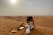 A woman dressed in white sitting in the desert beside a sandboard, gazing at the dunes.