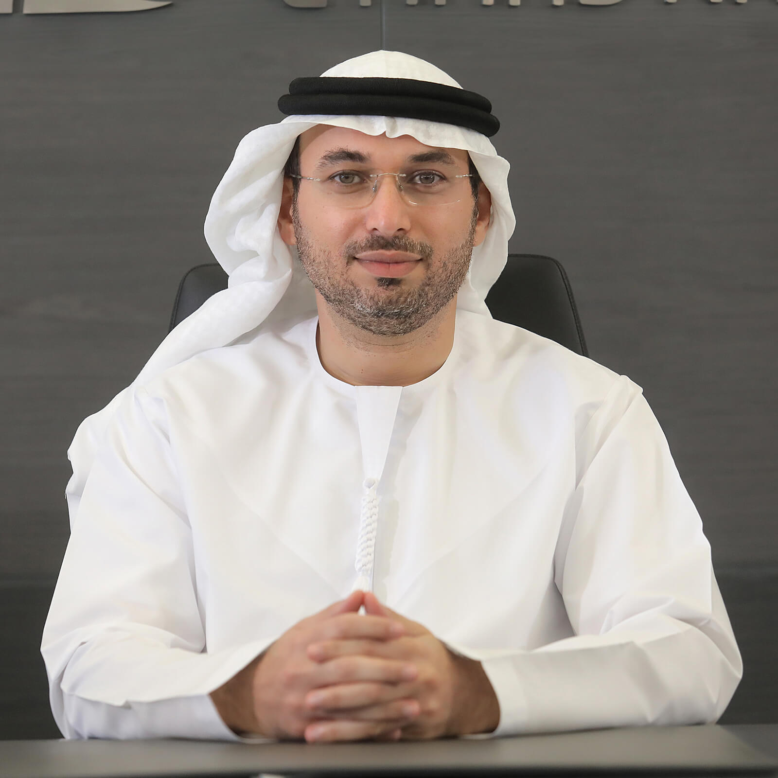 Portrait of an Emirati executive in a white kandura and black agal, seated at a desk with hands clasped, smiling confidently.