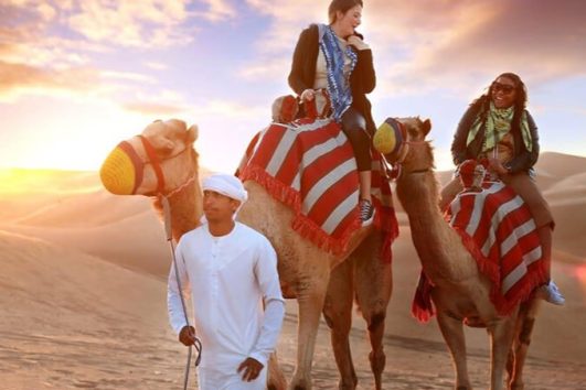 Two travelers on camels guided through the desert at sunset