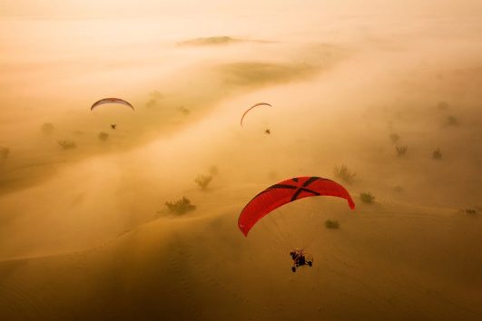 Multiple red paragliders flying over golden misty desert dunes during sunrise in Dubai