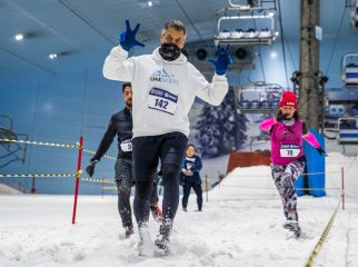 A man in a white hoodie with race number 142 running through deep snow at Ski Dubai, flanked by other competitors.
