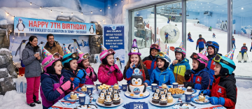 A group of children in winter jackets and party hats sitting at a table with penguin-themed cupcakes and a birthday cake.