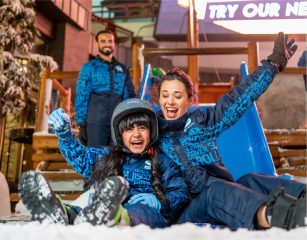 A laughing young girl and a woman sliding down a blue snow slide in matching navy blue winter gear at Ski Dubai.