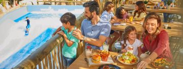 A family of four laughing and sharing a large plate of loaded nachos at a rustic wooden table in a vibrant restaurant setting.