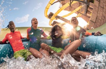 A family of four on a large blue raft screaming with excitement as they are splashed with water at the bottom of a yellow water slide.