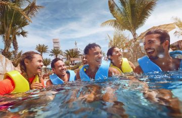 A diverse group of five friends wearing blue and yellow life jackets laughing while floating in a tropical pool surrounded by palm trees.