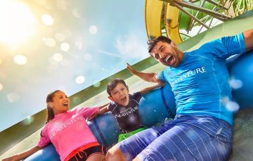 A close-up shot of a father, mother, and son screaming with joy as they descend a water slide on a blue raft.
