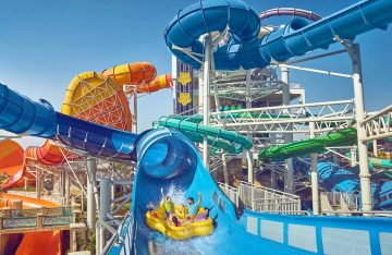 A wide shot of a massive water park structure with various blue, orange, and green winding slides and a yellow raft with riders.