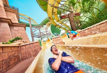 Two men racing down side-by-side yellow water slides with ecstatic expressions, surrounded by stone Mayan-style architecture and green slides.