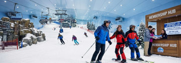 A group of skiers and snowboarders on an indoor ski slope next to a wooden Membership Hub counter at Ski Dubai.