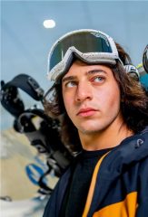A young man with long hair wearing ski goggles and carrying a snowboard, looking toward the slopes.