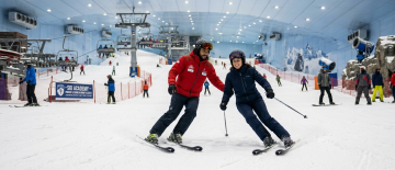 A professional ski instructor in a red jacket guiding a female student in a dark blue suit on the snow.