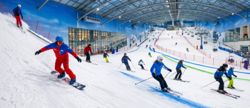Multiple skiers and a snowboarder in bright winter gear descending a wide, groomed indoor ski slope under a vaulted ceiling.