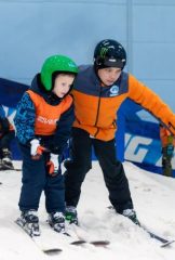 A professional ski instructor in an orange jacket guiding a young boy in a green helmet during a ski lesson.
