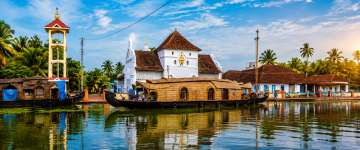 Traditional Kerala houseboats near a historic church and waterfront in Fort Kochi surrounded by coconut palms.