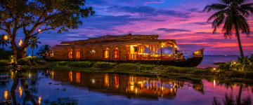 Luxury Kerala houseboat illuminated at sunset on the peaceful Kumarakom backwaters surrounded by tropical palms.