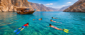 Guests snorkeling in crystal-clear waters beside a traditional dhow in the Musandam fjords.